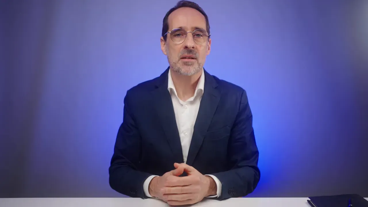 Referent vor blauem Hintergrund, ruhig sitzend mit gefalteten Händen auf dem Tisch